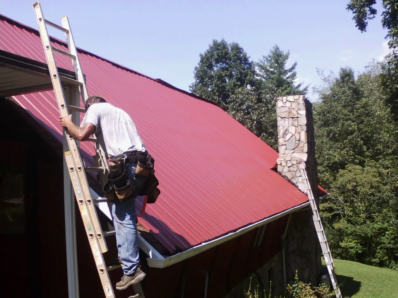Roofer on a ladder beside a red metal roof and stone chimney — Soffit & Fascia Repair by Roofing Brothers in West Baldwin
